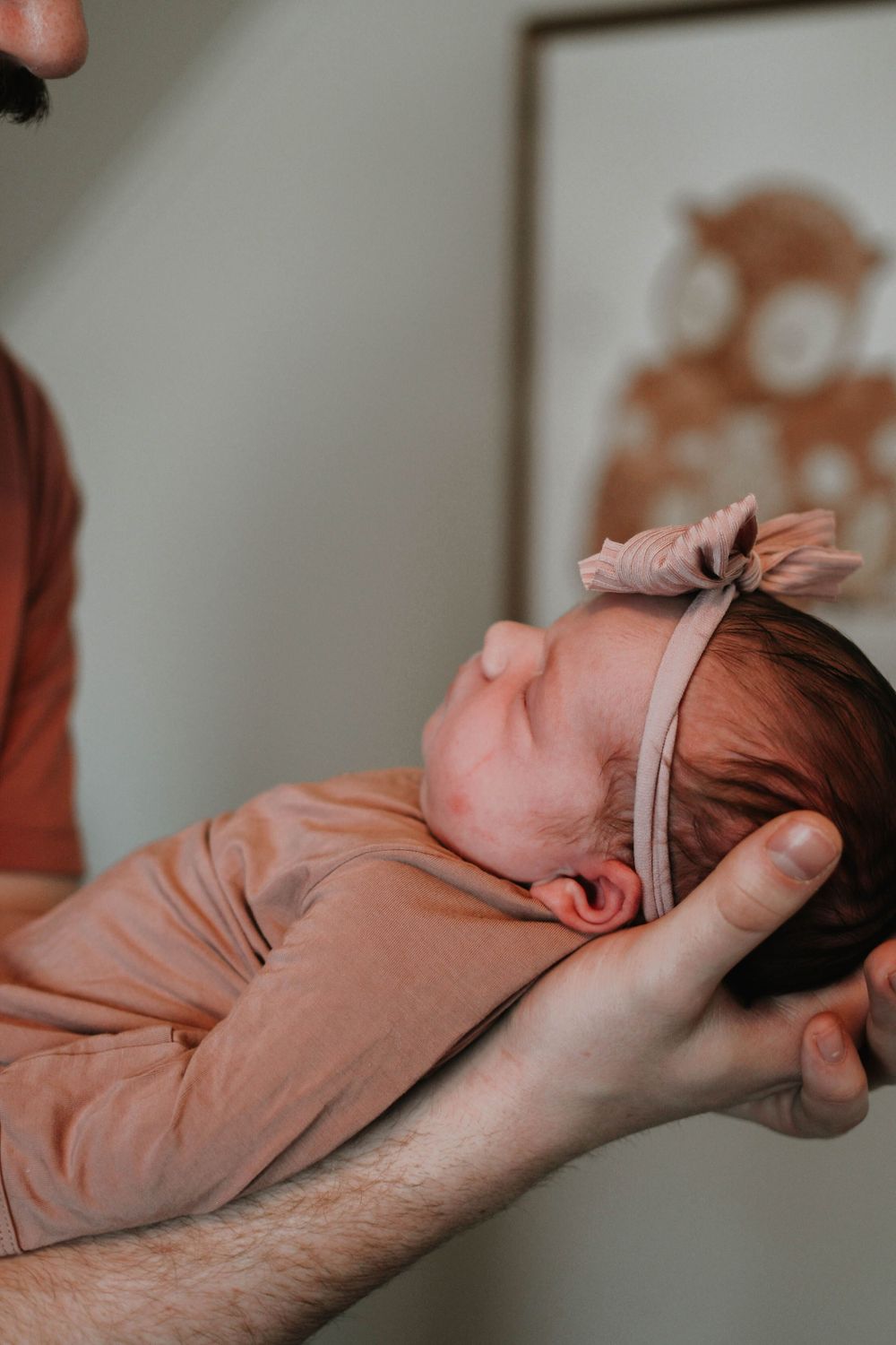 A sleeping newborn baby wearing a peach-colored onesie and headband is cradled gently in someone's hands.