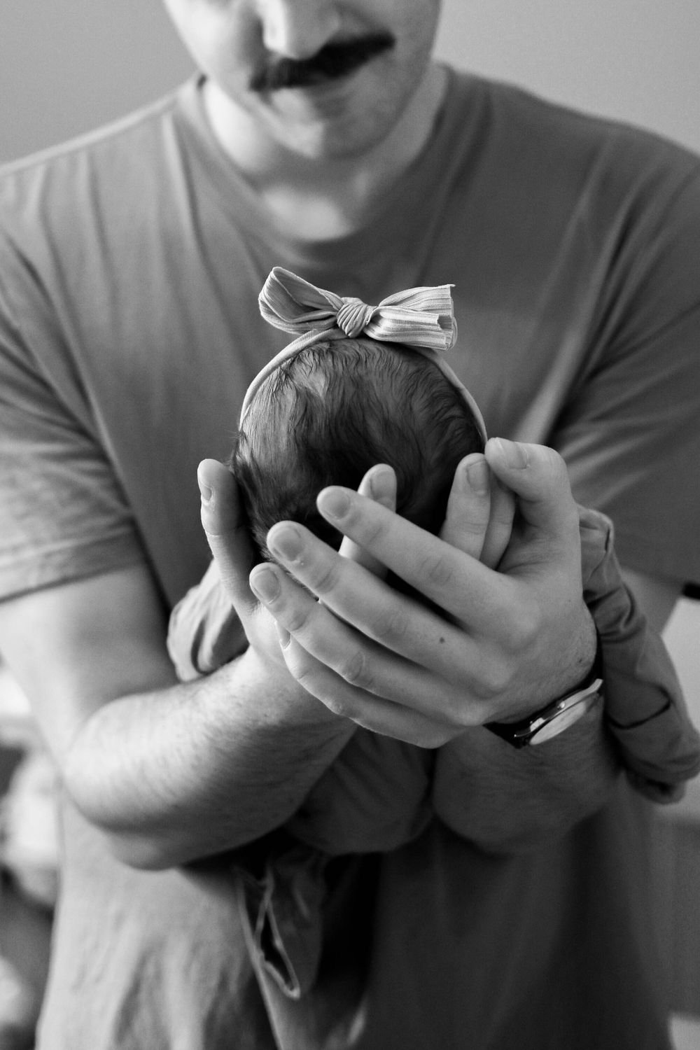 A newborn baby wearing a white bow headband being gently cradled in hands.