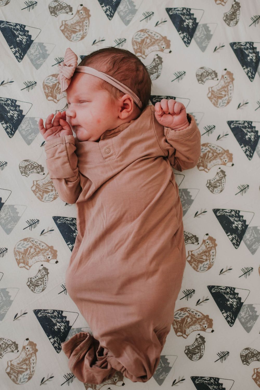 Sleeping newborn in pink headband and tan swaddle lying on patterned triangle print blanket.