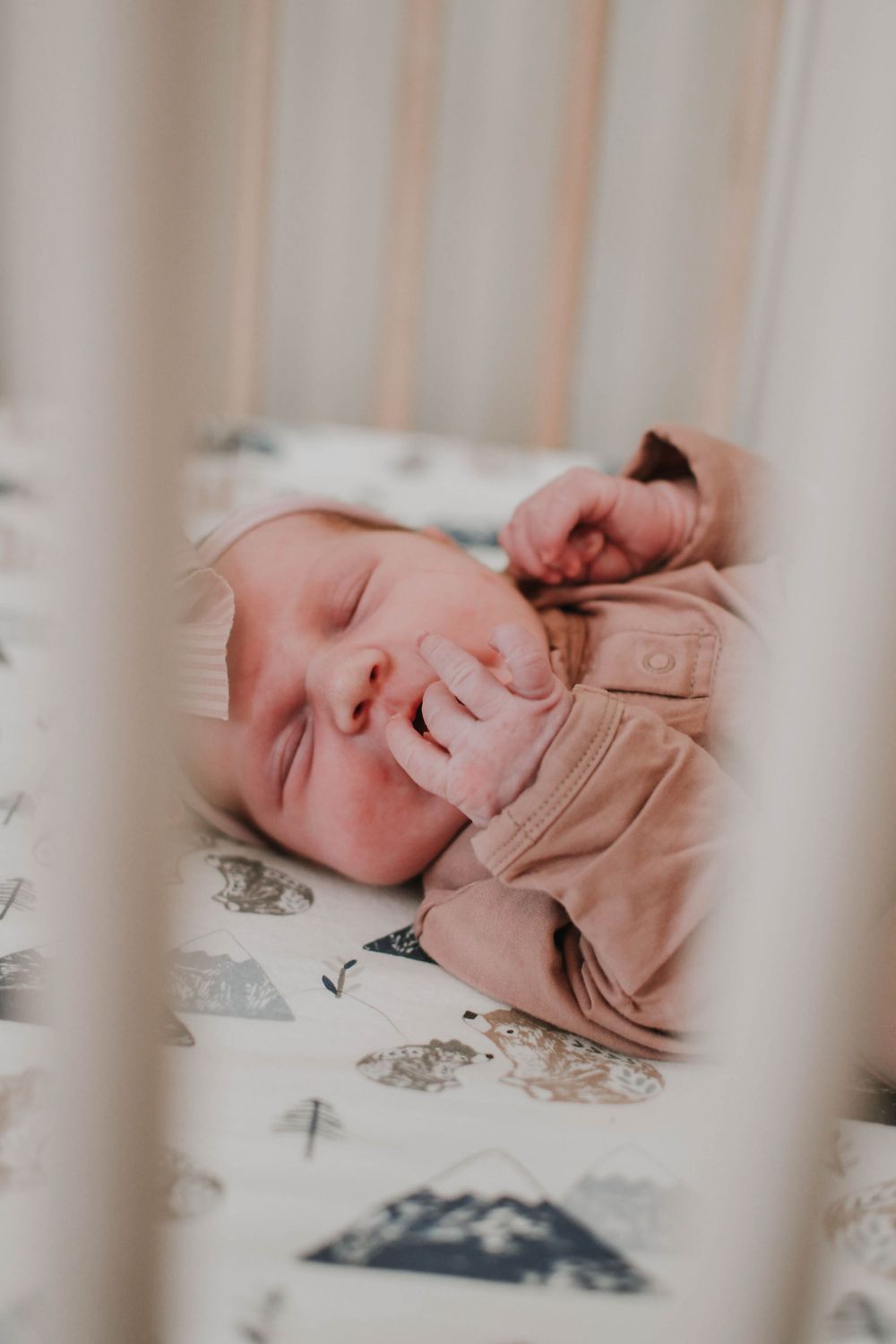A peaceful sleeping baby in a light brown outfit lies in a crib with patterned bedding.
