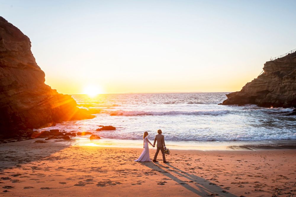 Two silhouettes walking hand in hand on a beach at sunset with dramatic rocky cliffs and ocean waves.