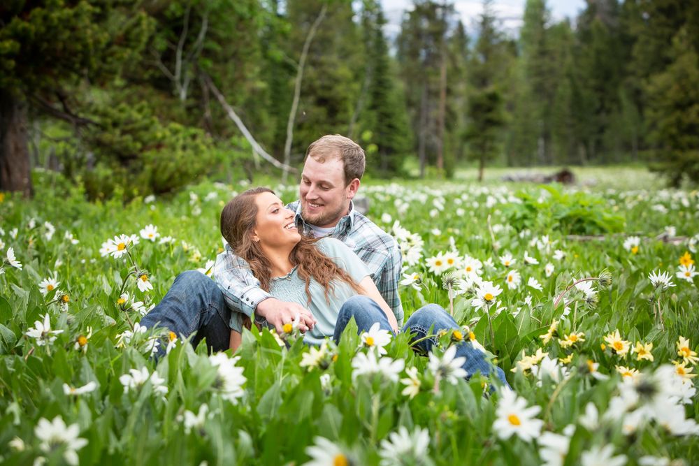 A couple shares a lighthearted moment while sitting in a mountain meadow filled with blooming white wildflowers.