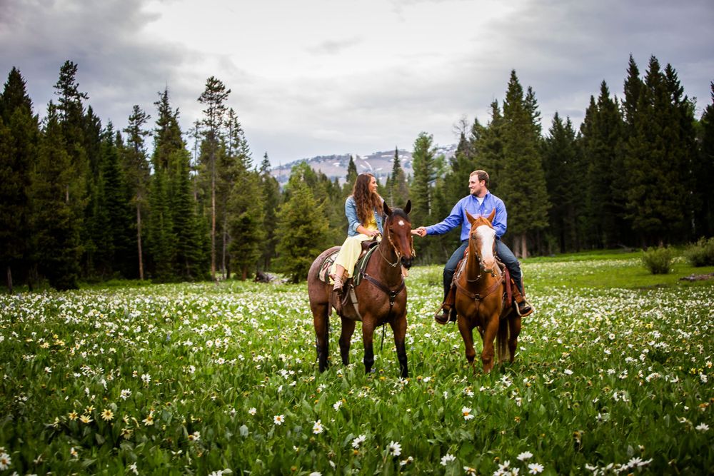 Two riders on horseback in a mountain meadow filled with wildflowers and surrounded by evergreen forest.