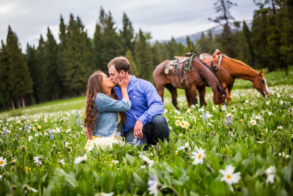 A couple sits in a meadow of wildflowers while two horses graze in the background against a mountain forest landscape.