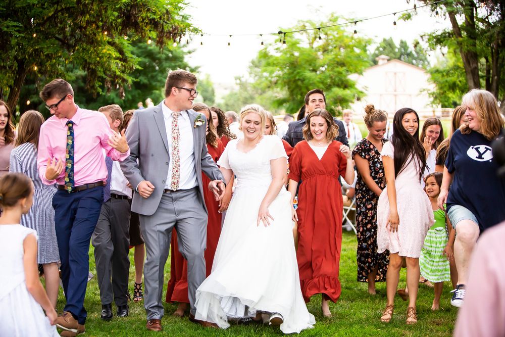 A joyful wedding party in formal attire walks together outdoors on a sunny summer day surrounded by greenery.