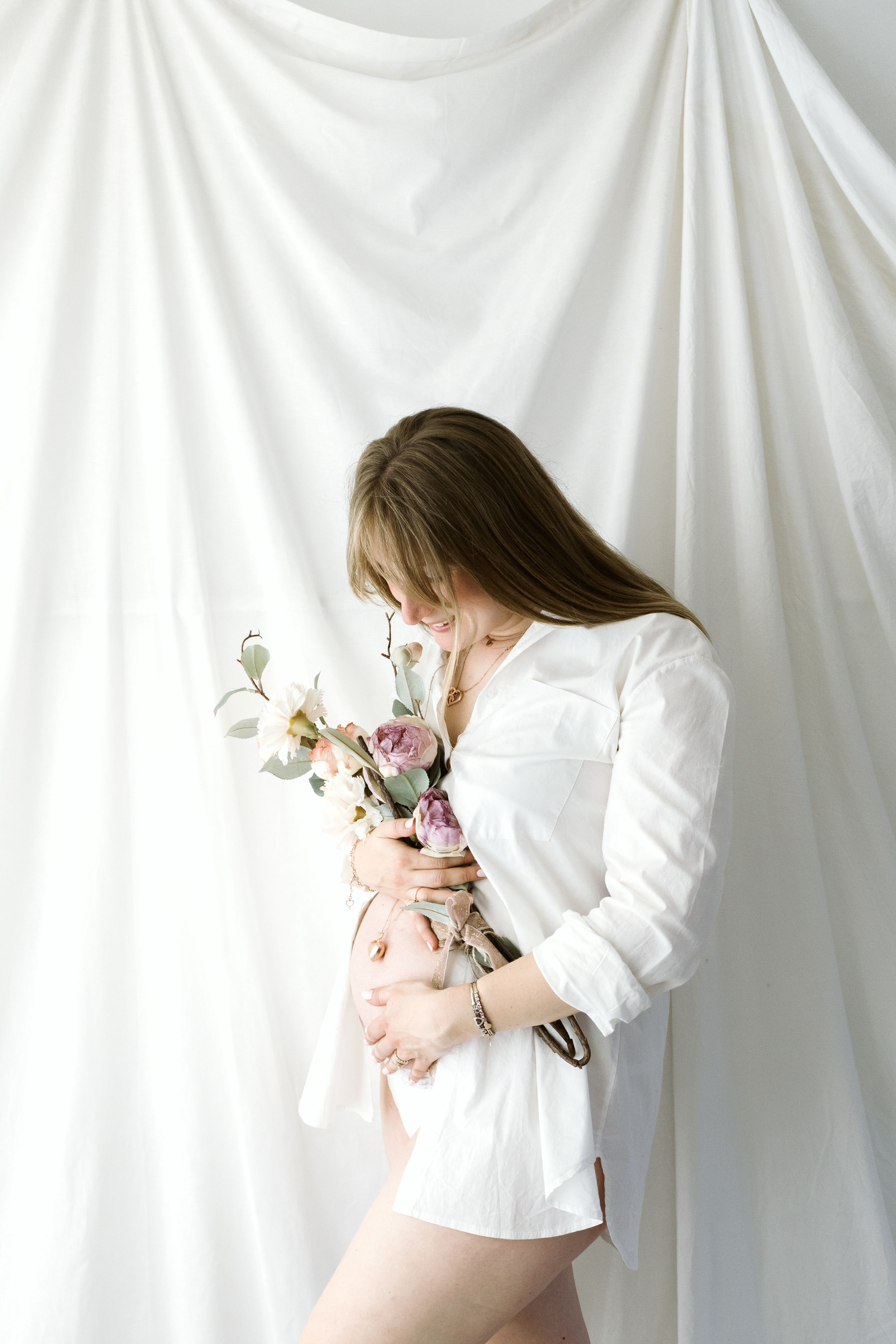 Donna incinta in camicia bianca che tiene delicati fiori rosa e bianchi su sfondo bianco. Servizio Maternità Cassine AL.