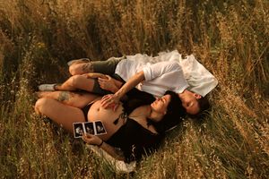 Coppia sdraiata insieme sull'erba alta durante un romantico servizio fotografico maternità all'aperto al tramonto.