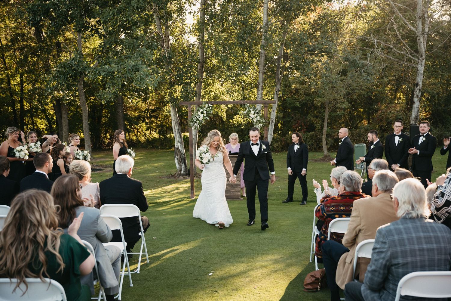 Outdoor wedding ceremony in progress with guests seated on white chairs among tall trees.