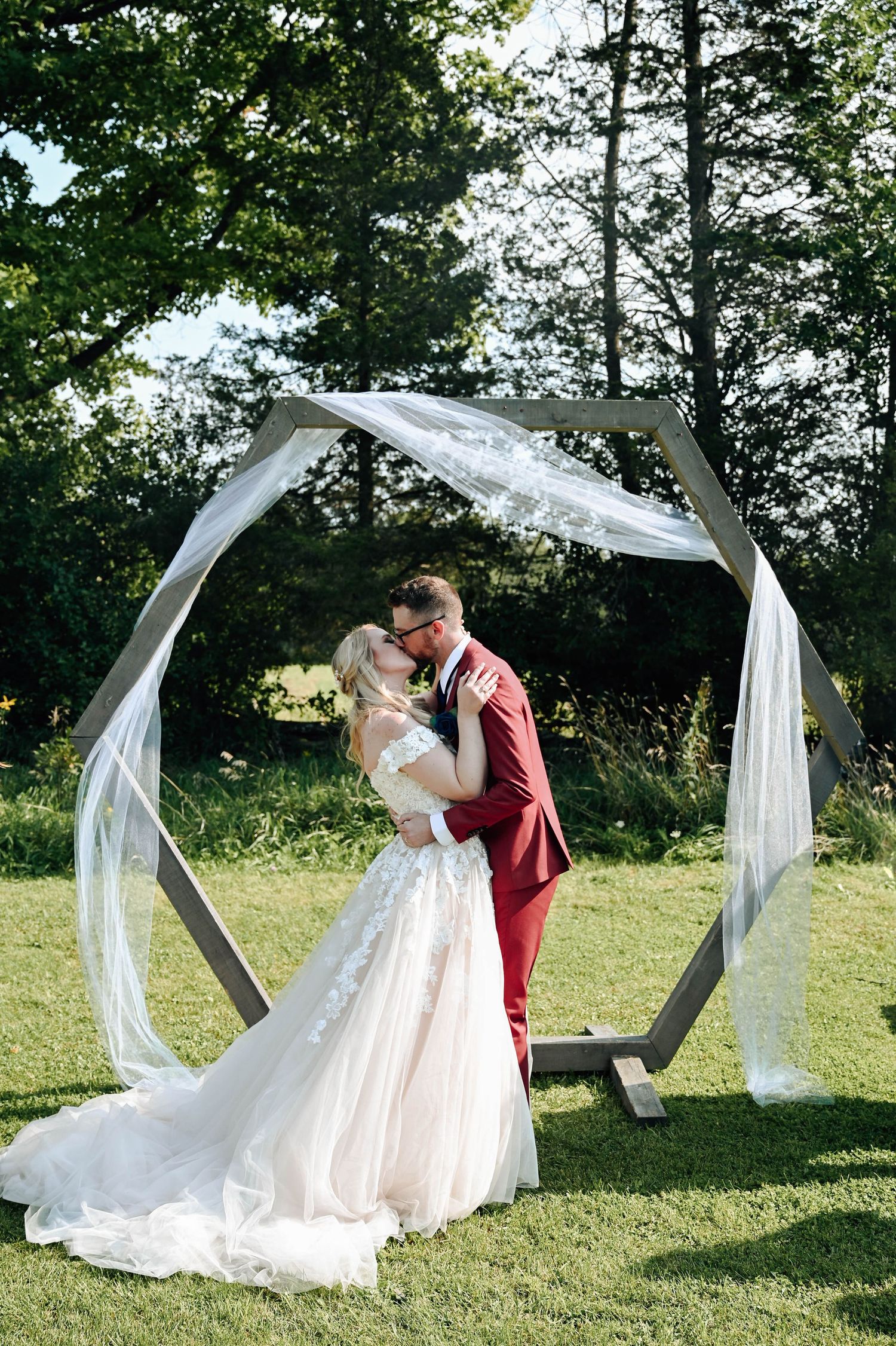 Couple shares romantic kiss under hexagonal wedding arch draped with white fabric on sunny garden lawn.