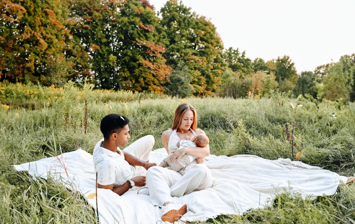 People relax on white blanket in summer meadow with tall grass and autumn trees in background.