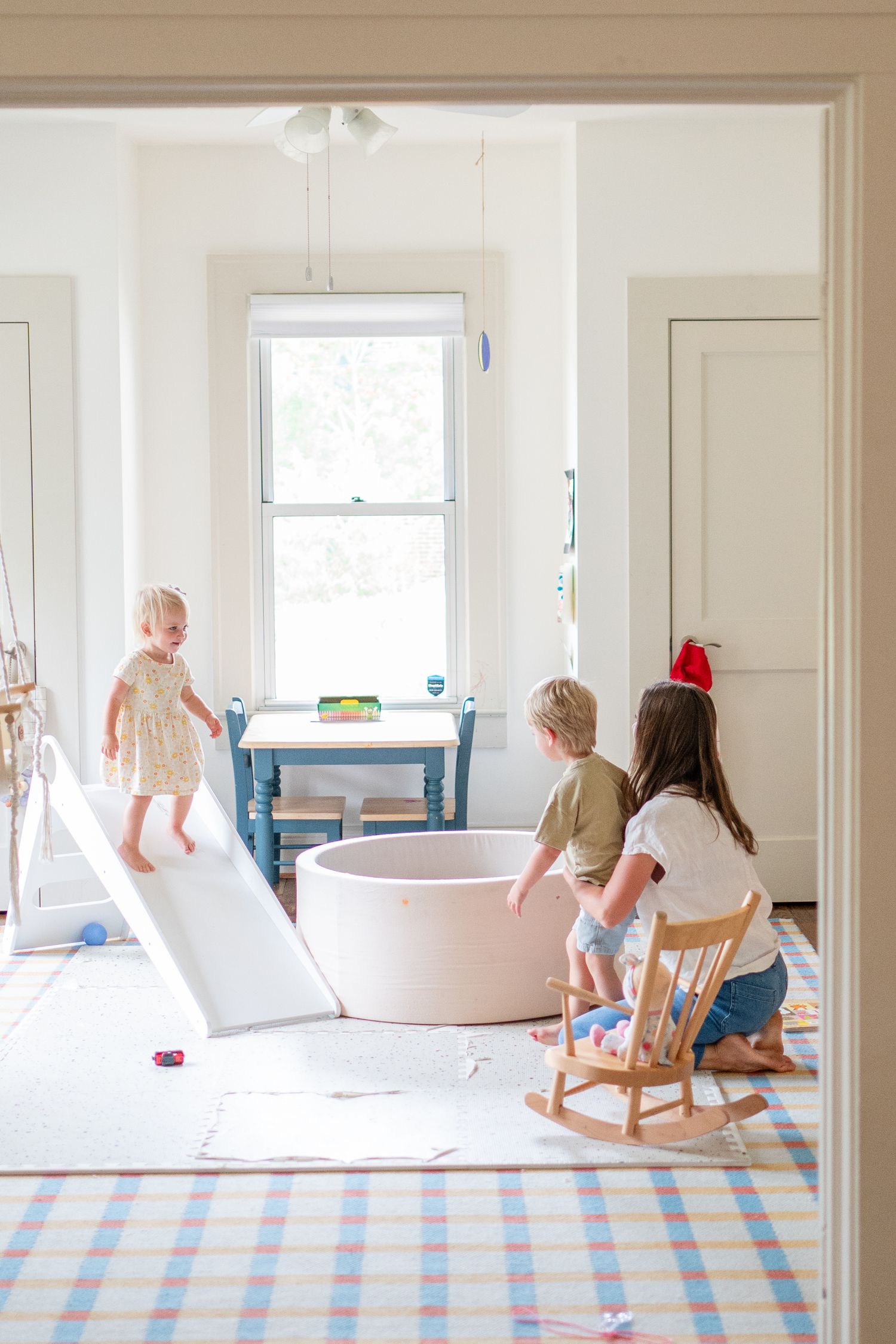 Children playing on a slide with a white soaking tub in a bright interior room with a blue checkered rug.