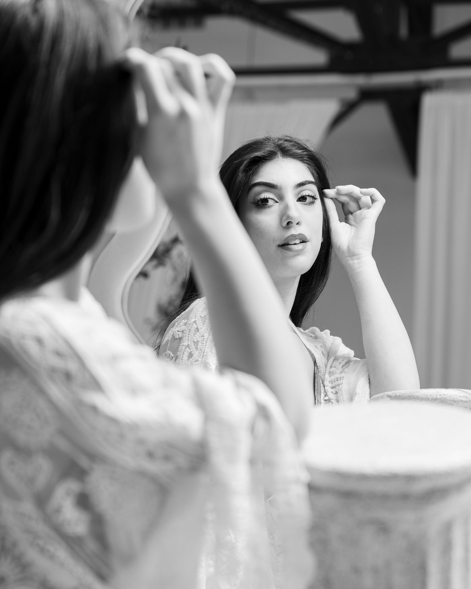 Black and white image of someone applying makeup while looking in a mirror wearing an elegant lace top.