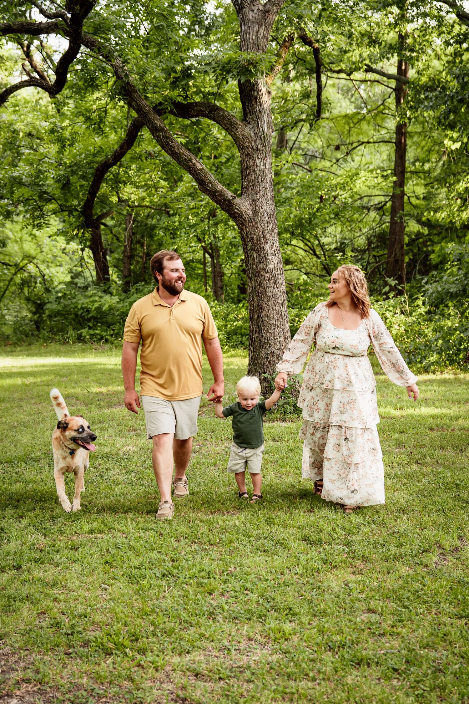 Family walking with dog through wooded park on sunny day.