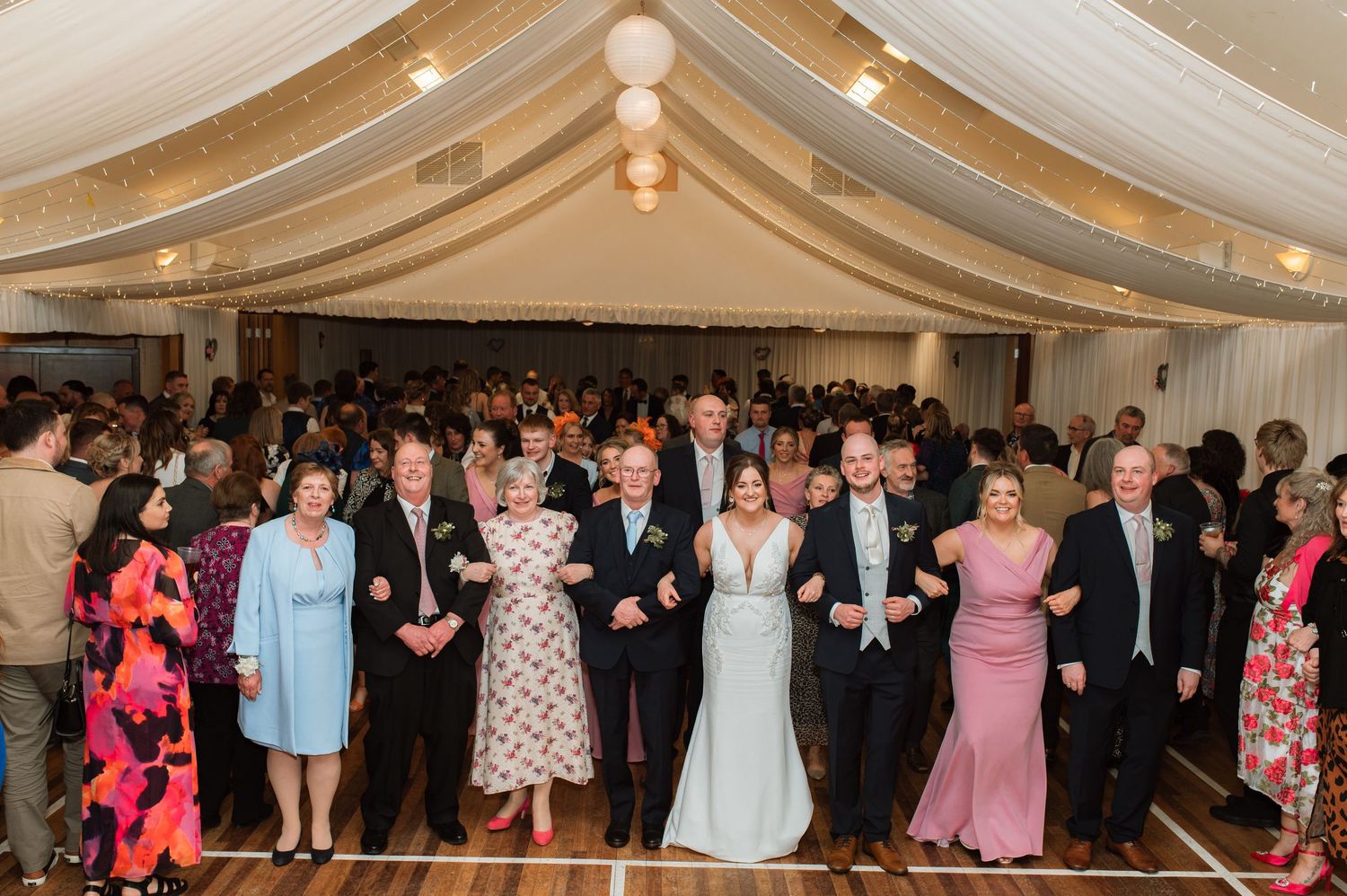 Wedding reception guests gathered in an elegant tent-like venue with draped fabric ceiling and string lights for dancing.
