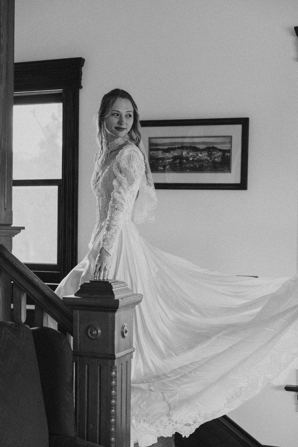 Vintage-inspired black and white photograph of a bride in a flowing lace wedding dress standing on a wooden staircase.