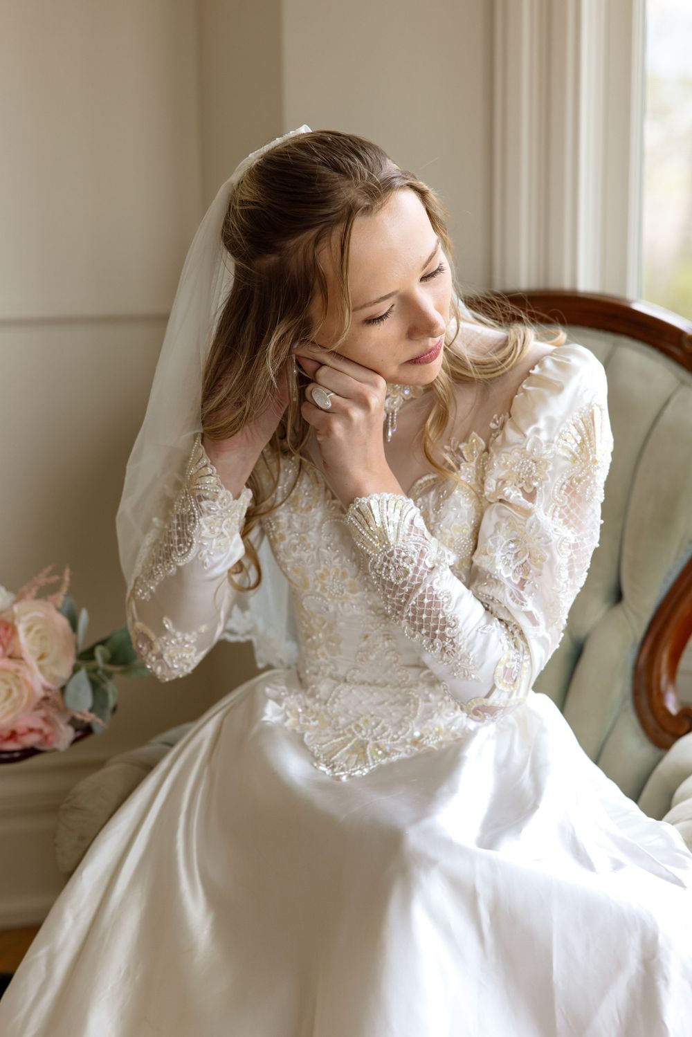 Wedding bride in long sleeve lace gown sits on vintage chair while using mobile phone near window in natural light.