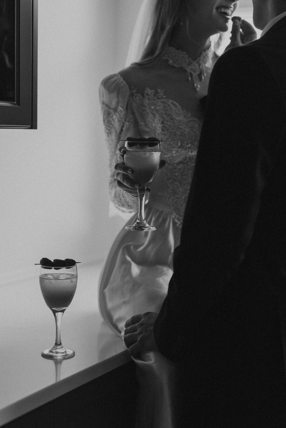 Black and white photo of a couple at a formal event with wine glasses, showing elegant lace dress details.
