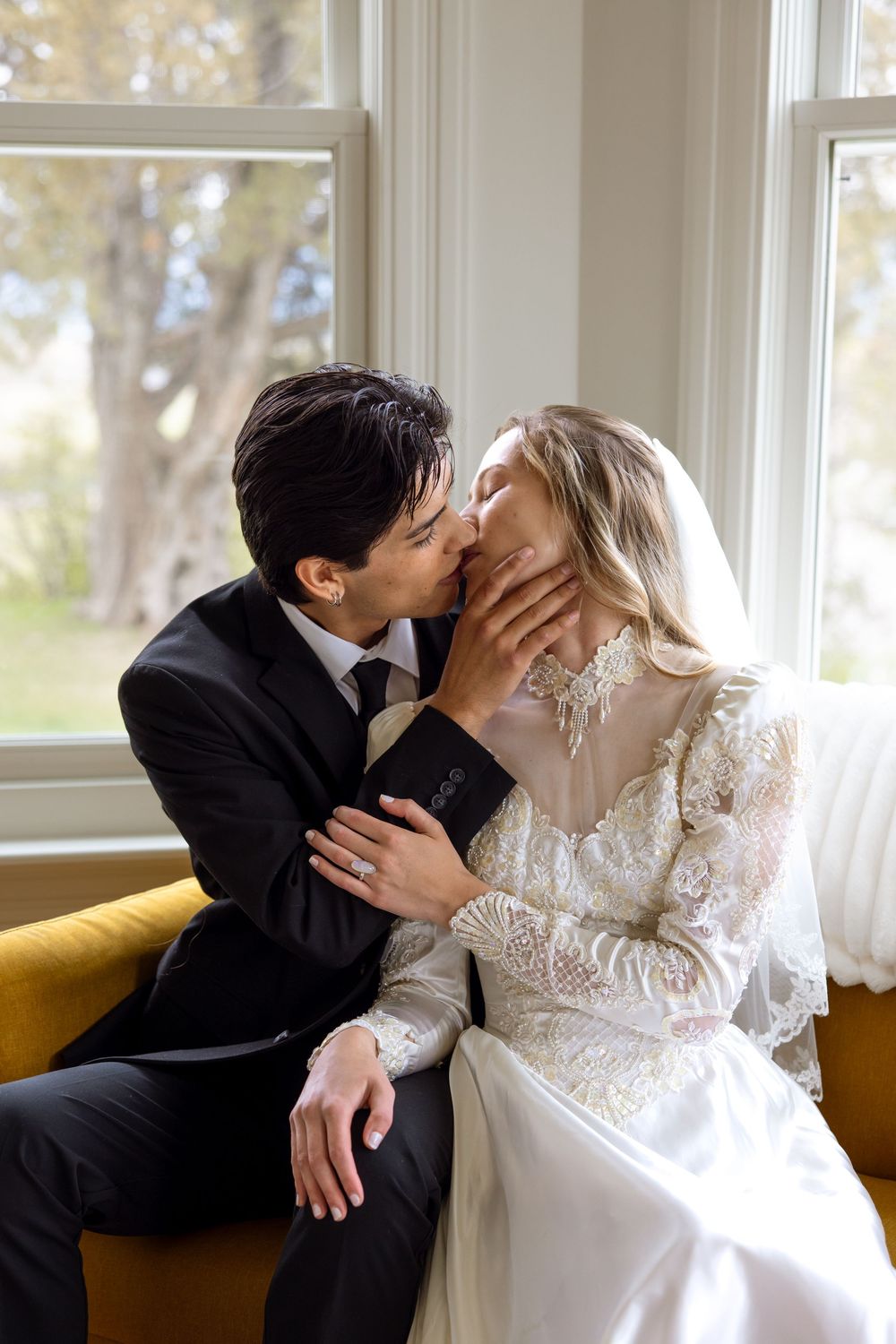 A romantic wedding couple shares a tender kiss while seated by sunlit windows on a yellow couch.