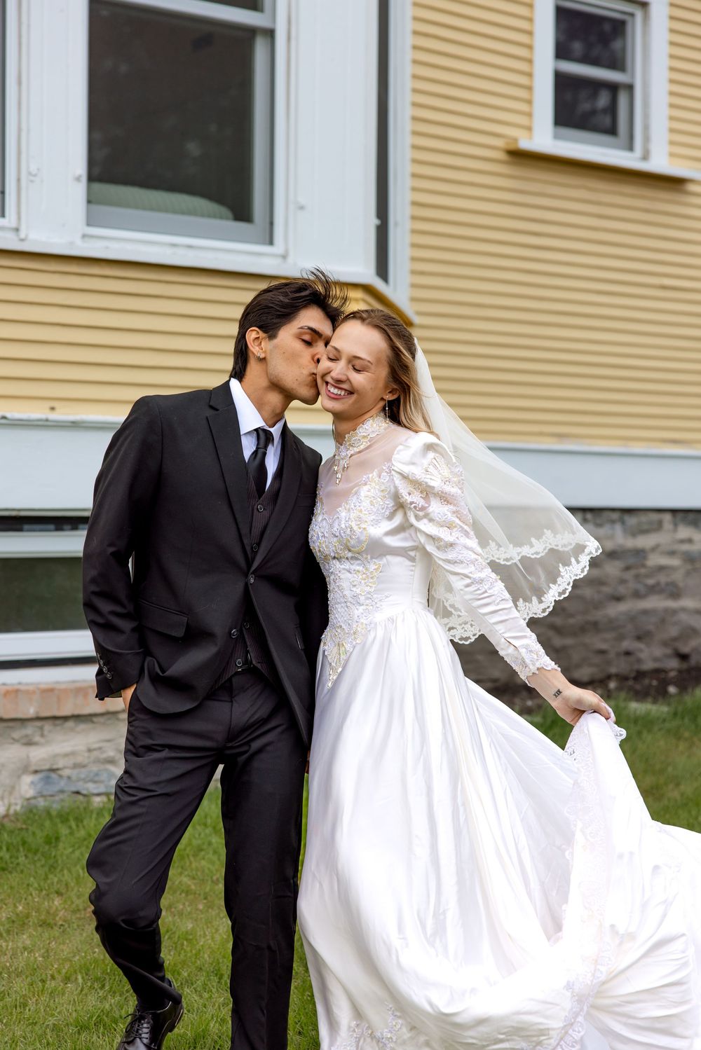 A couple embrace outdoors on their wedding day, with the bride in a white lace dress and groom in a black suit.