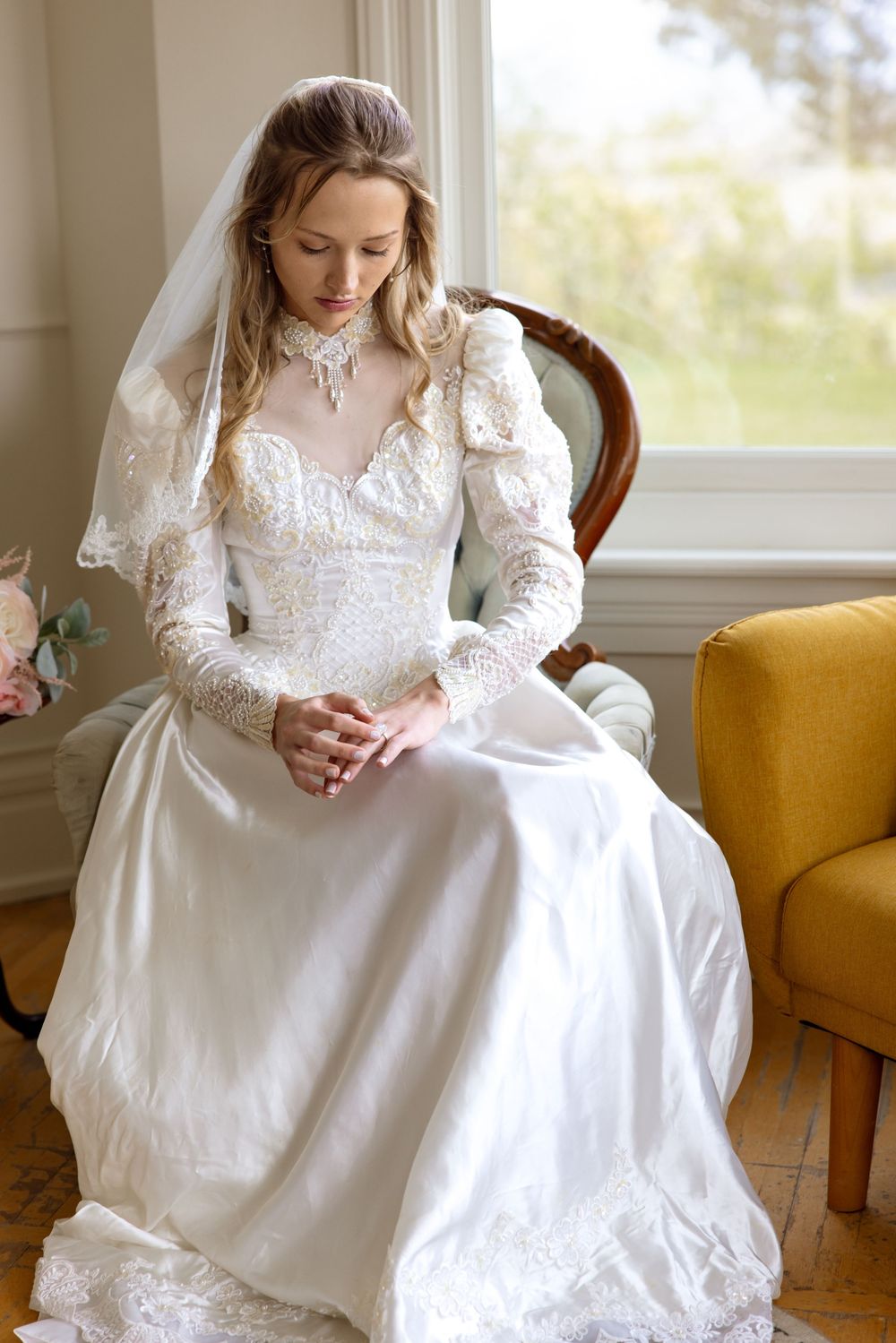 A bride in a vintage-style lace wedding dress with long sleeves sits thoughtfully by a window in a room with a yellow chair.