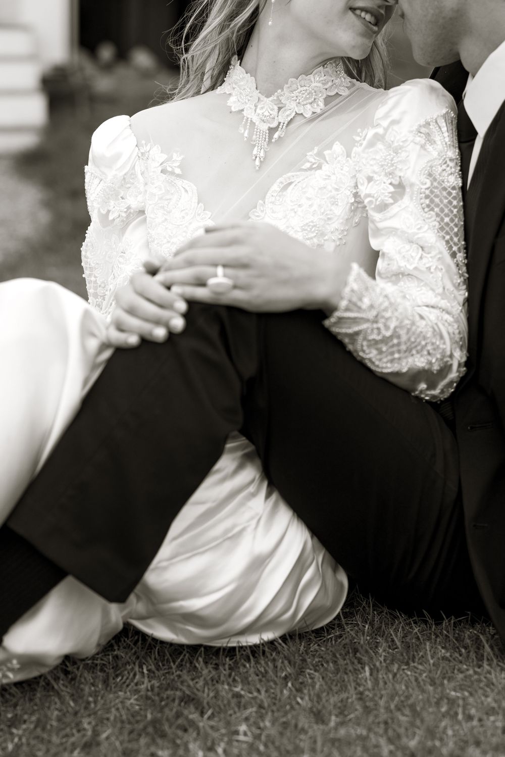 Black and white closeup of a romantic wedding moment showing vintage lace sleeves and formal attire while sitting on grass.
