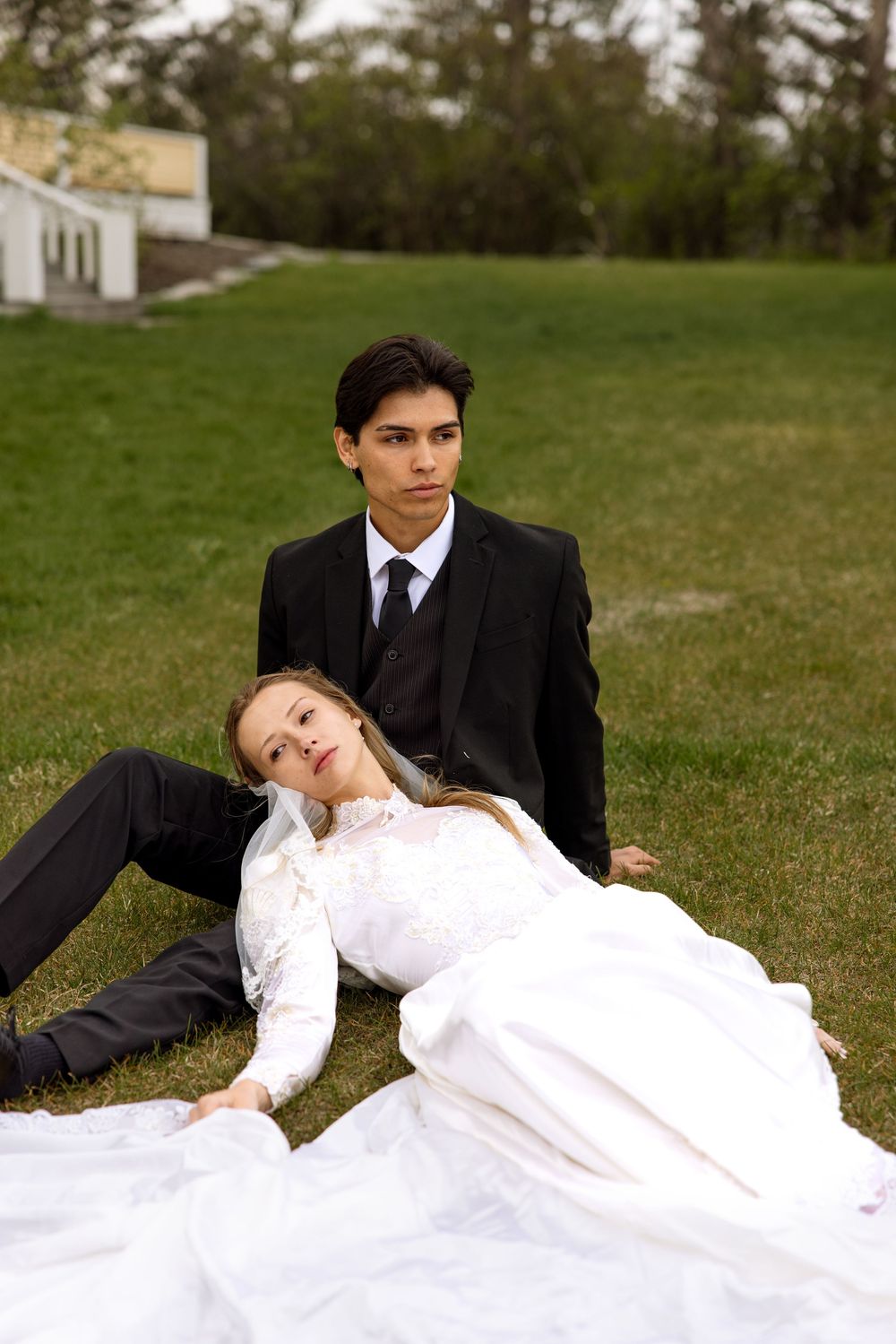 A couple in formal wedding attire relaxes on a grassy lawn in a romantic outdoor portrait.