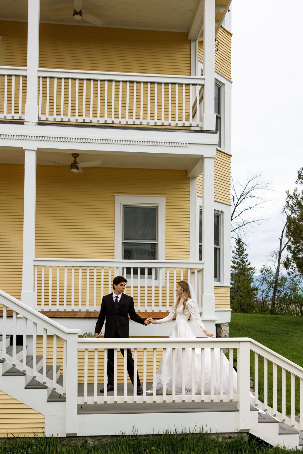 A bride and groom holding hands on the white porch steps of a yellow Victorian style house.