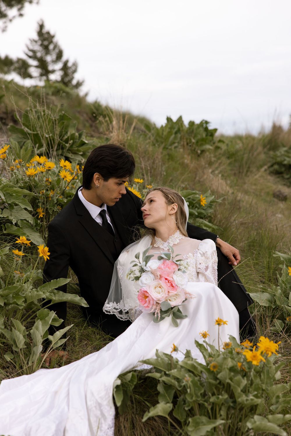 A couple in wedding attire sits among yellow wildflowers on a hillside, holding a bouquet of pink roses.