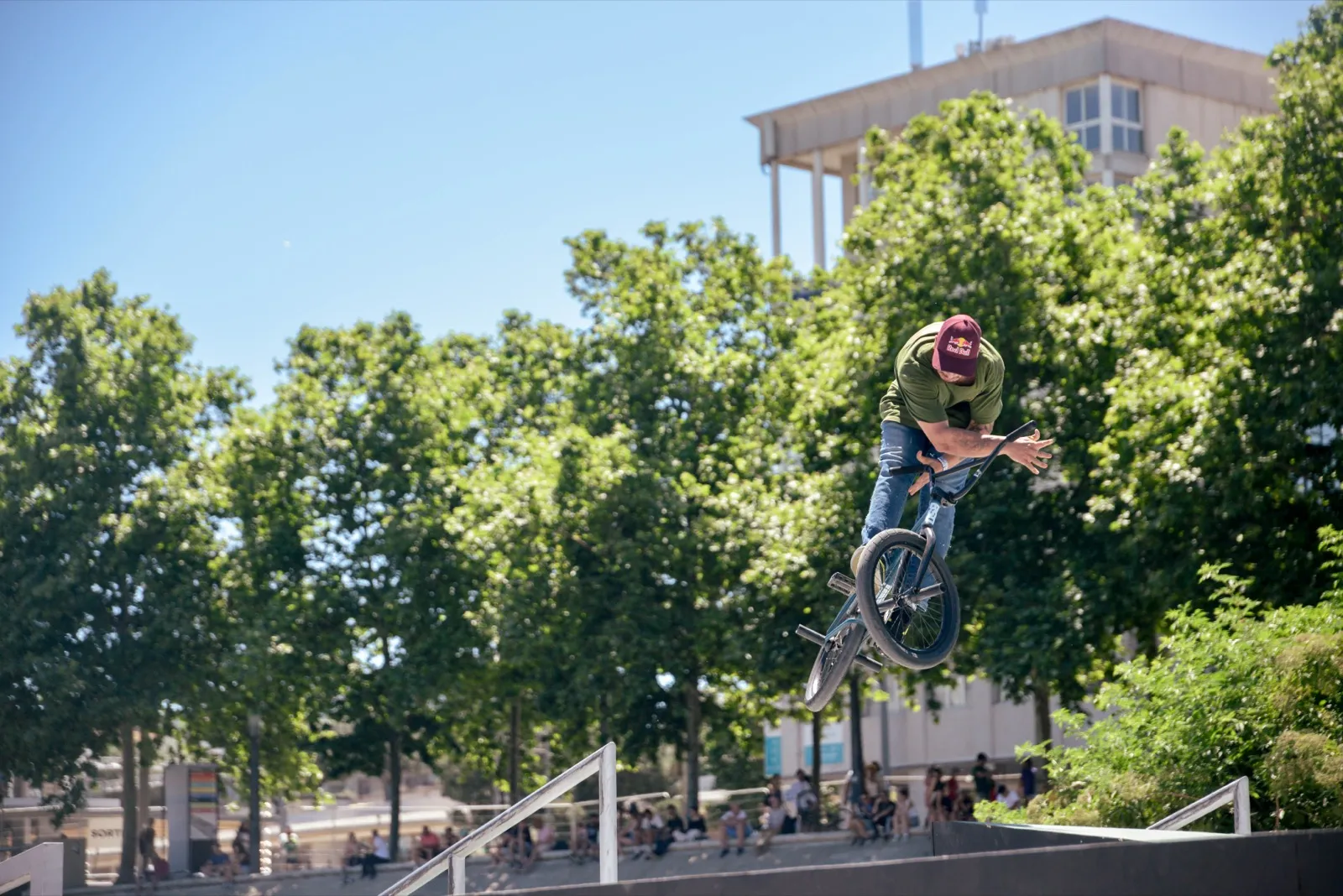 Rider BMX dans les airs au-dessus d’un skatepark, arbres et bâtiments en fond.