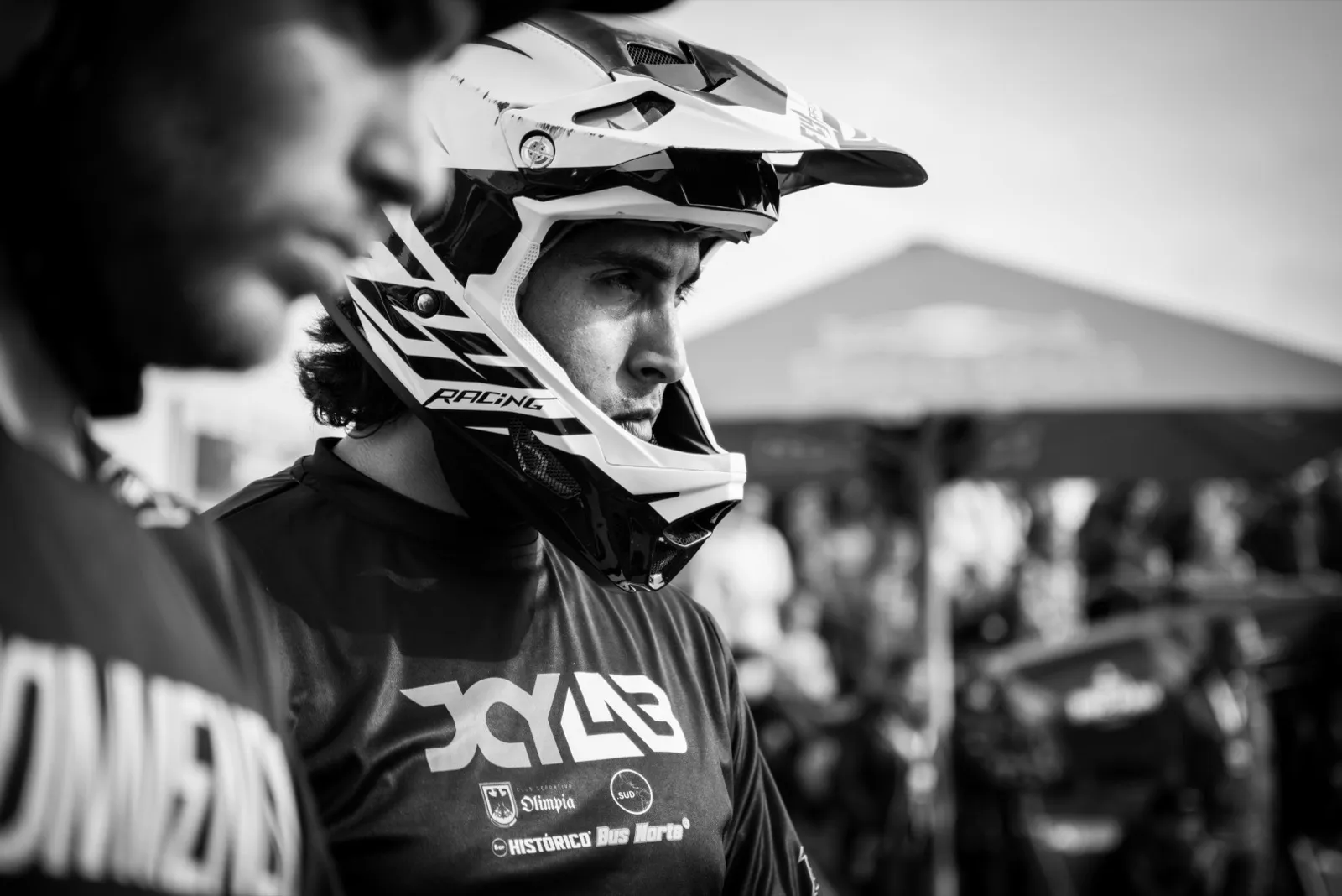Portrait en noir et blanc d’un pilote de VTT en tenue FOX et casque intégral, observant la scène, foule floue en arrière-plan.