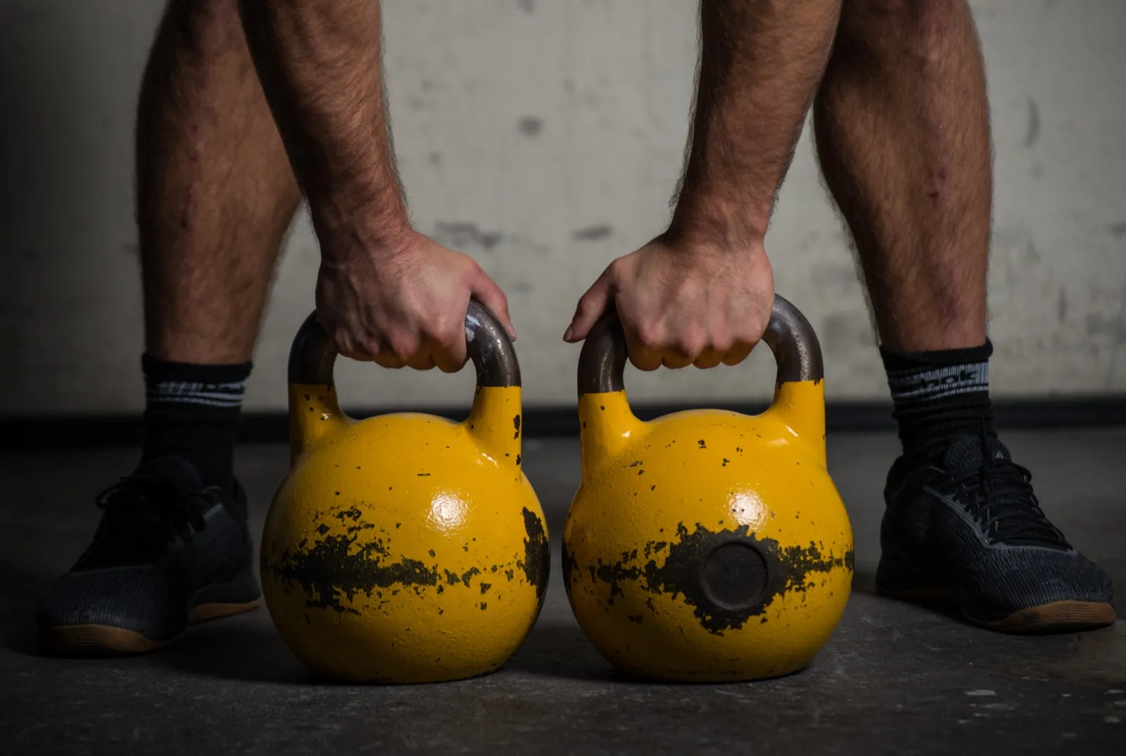 Plan serré sur les jambes d’un athlète tenant deux kettlebells jaunes posés au sol, prêt pour un exercice de force.