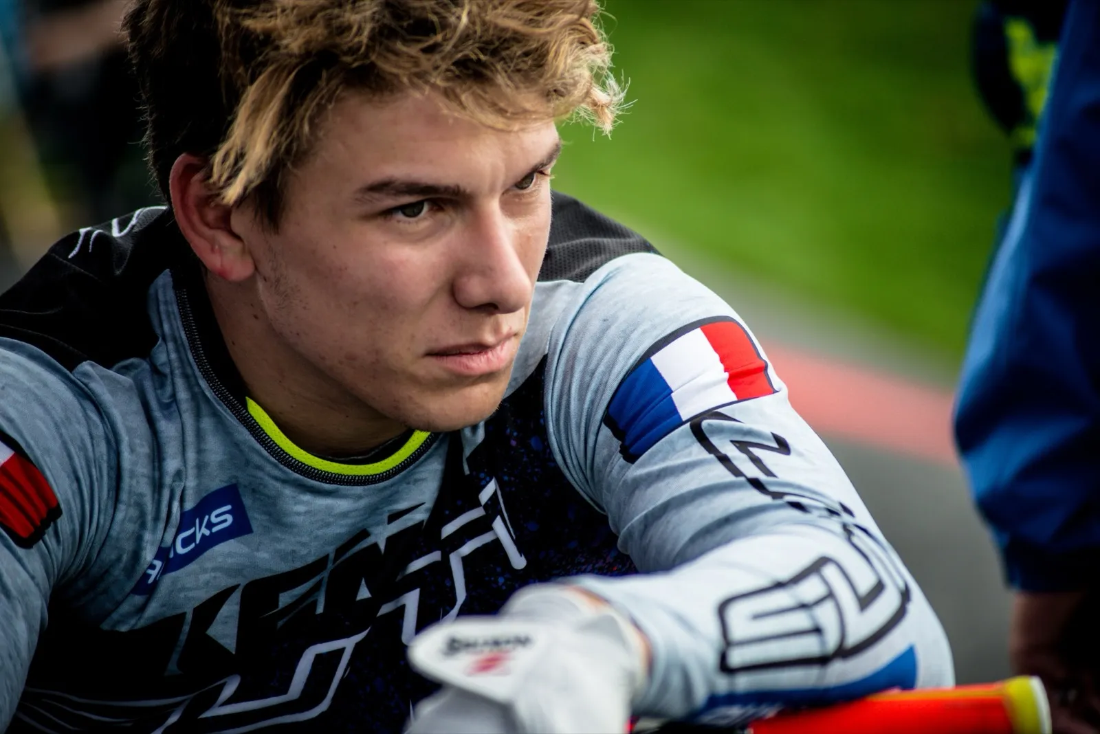 Plan serré sur un rider BMX français, appuyé sur son guidon, regard déterminé avant la course.