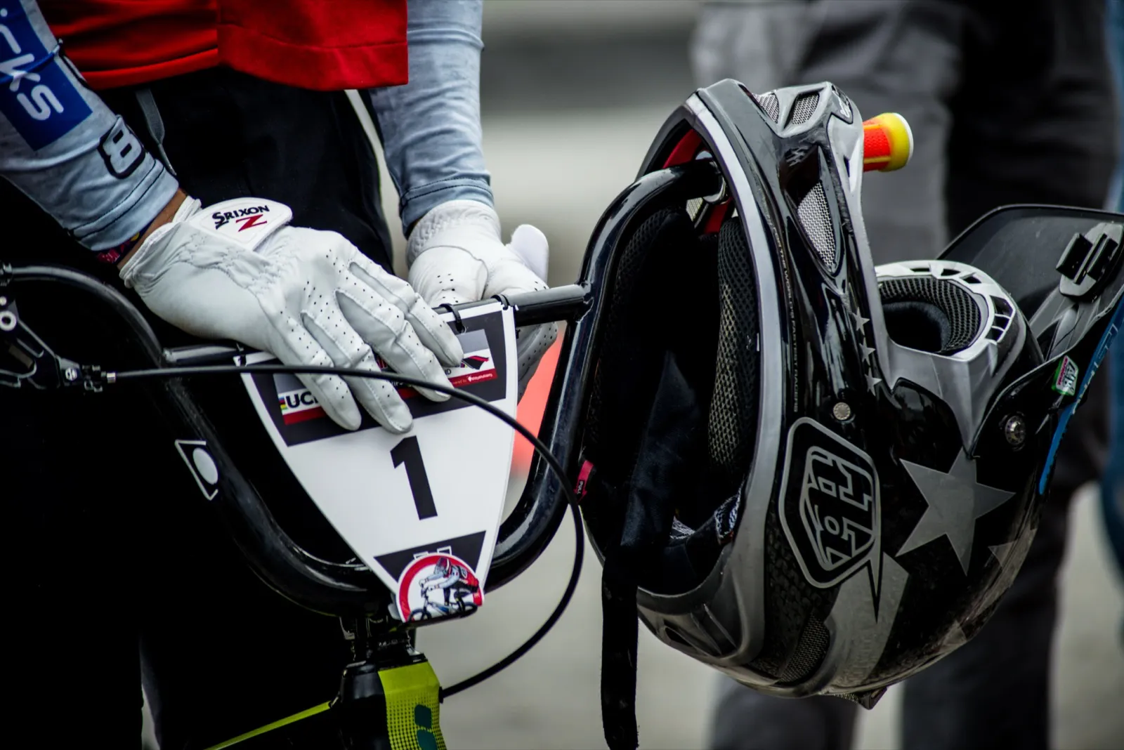 Gros plan sur un rider BMX tenant son casque et son guidon, gants blancs et numéro de plaque visible.