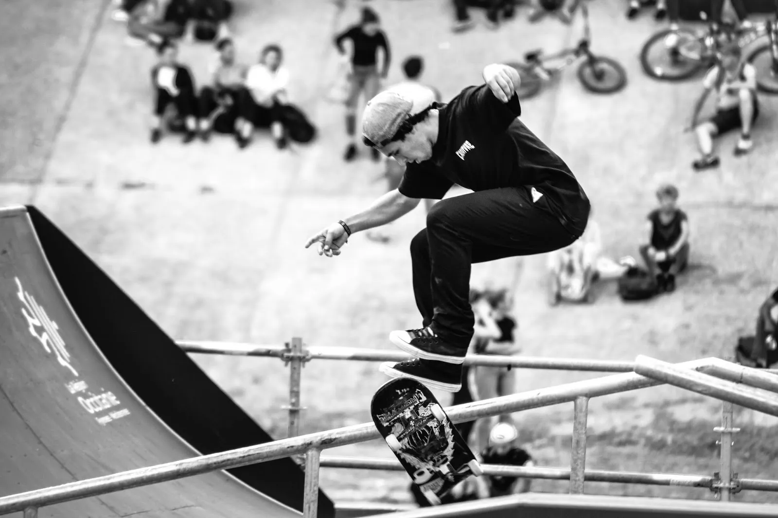 Skateur effectuant un trick sur une barre en descente, photo en noir et blanc avec foule en fond.