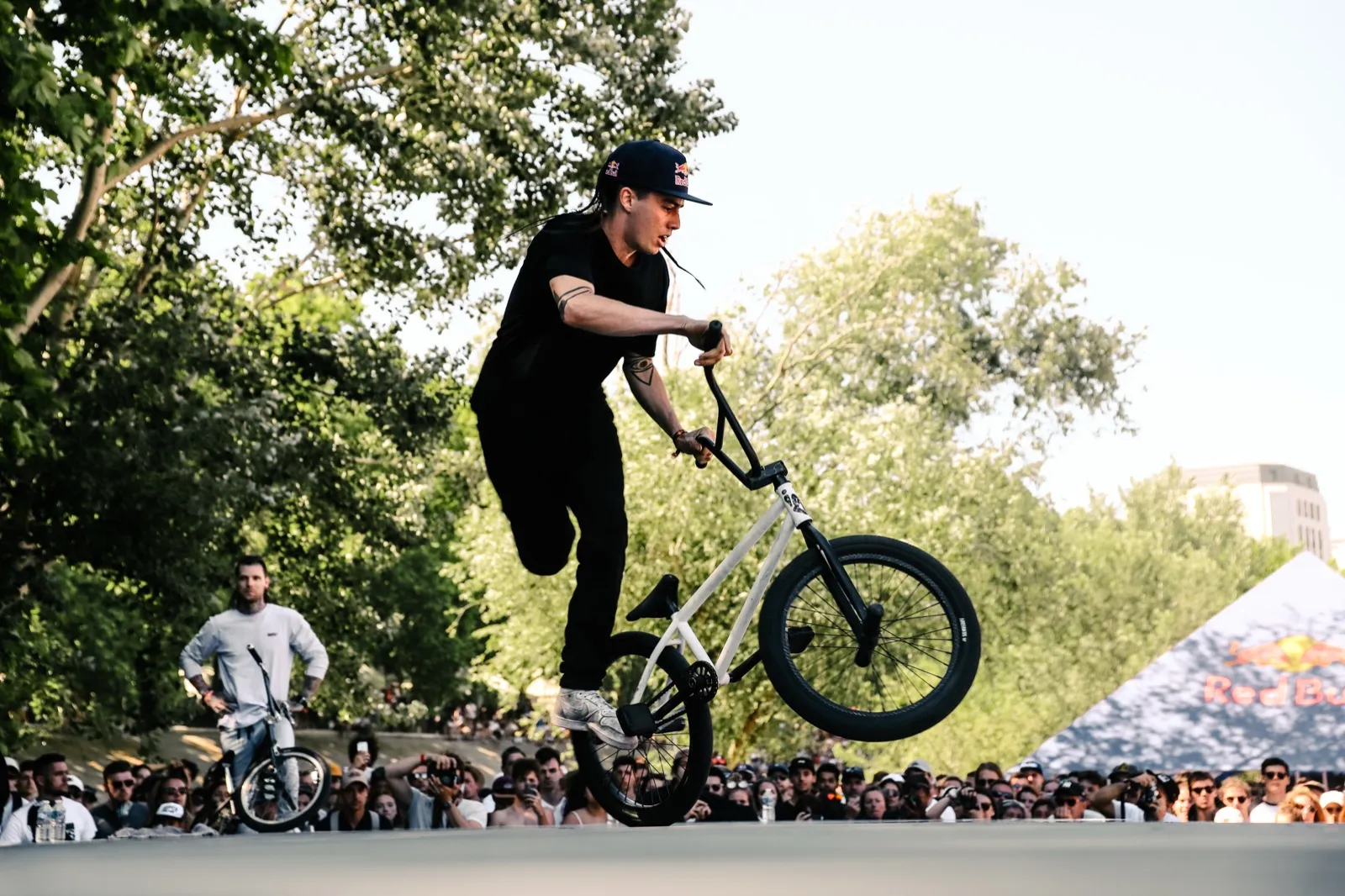 Figure de BMX flat en plein saut, foule rassemblée en fond sous les arbres.