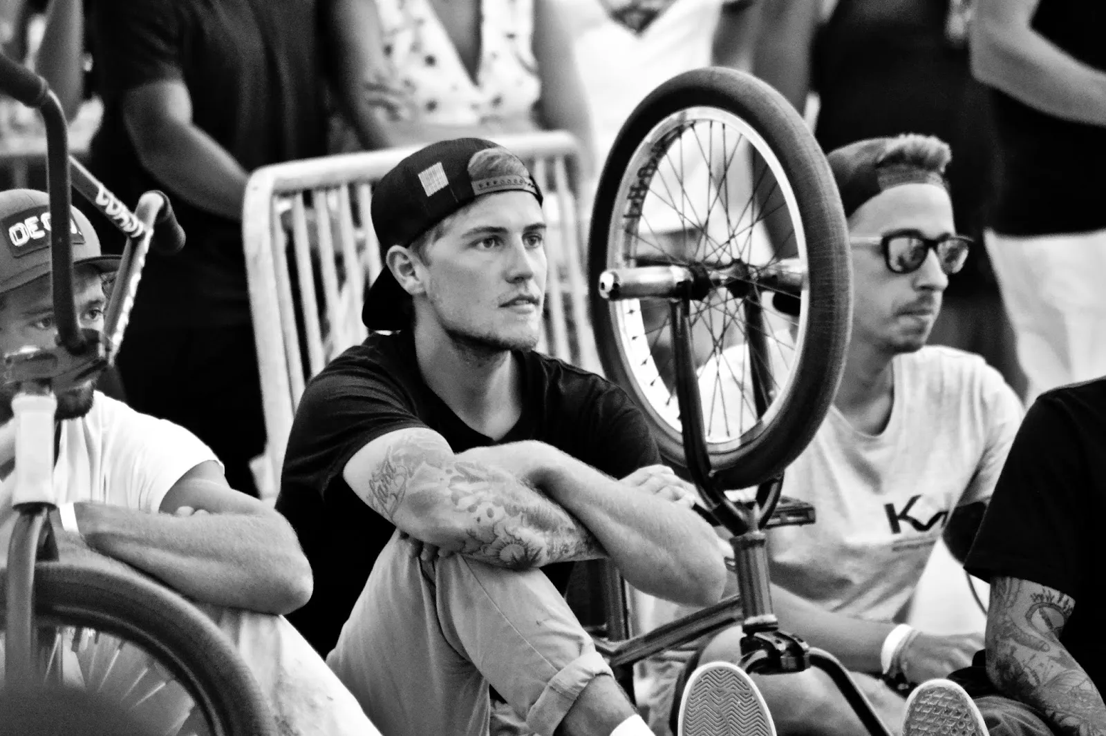 Scène en noir et blanc d’un rider BMX assis au sol entouré d’autres jeunes, avec un vélo retourné au premier plan.