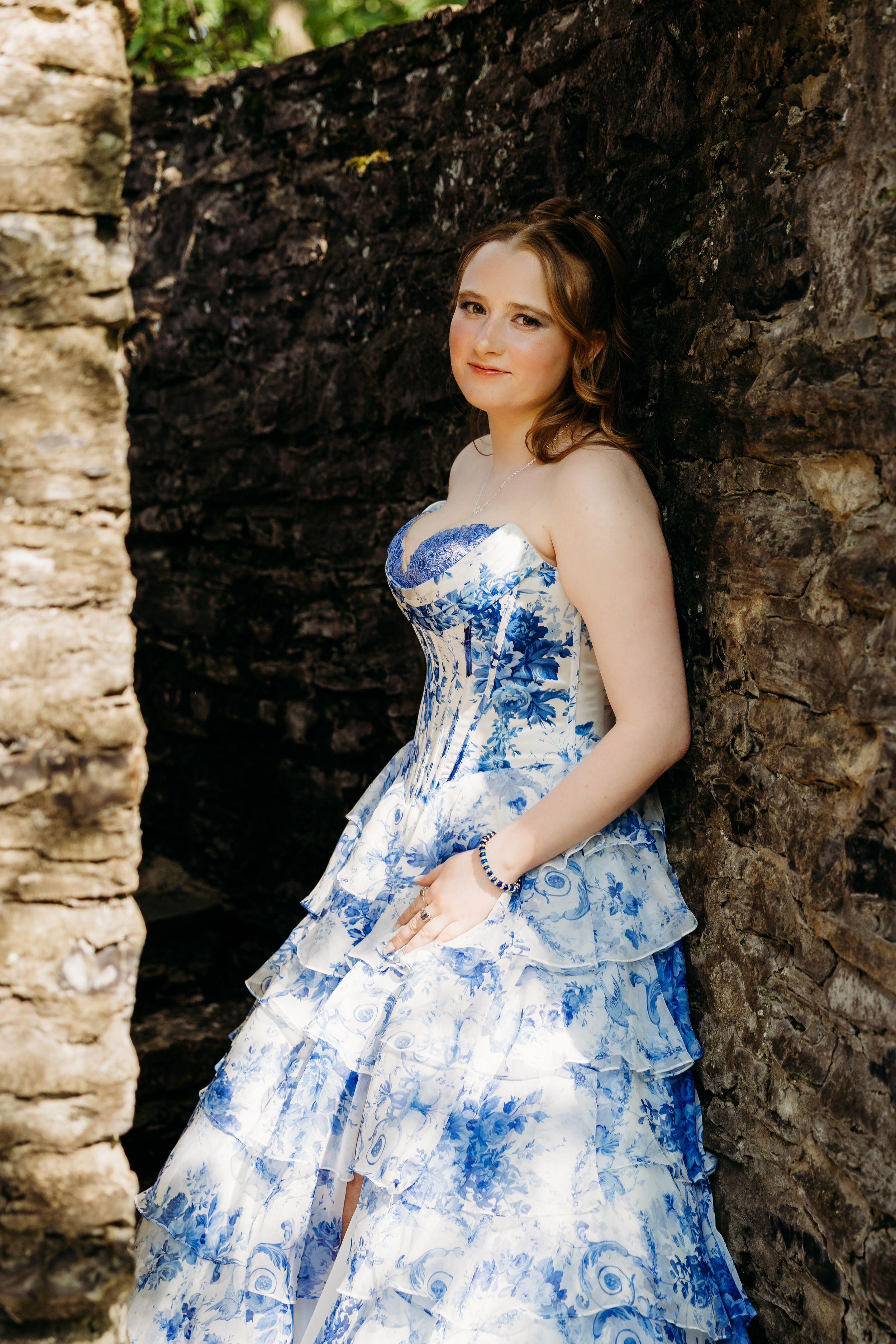 A strapless blue and white floral ball gown with a corset bodice photographed against a stone wall.