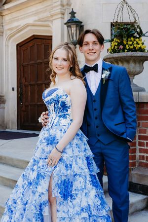 A couple in formal wear poses together, with a vibrant blue and white floral ball gown and navy blue tuxedo.