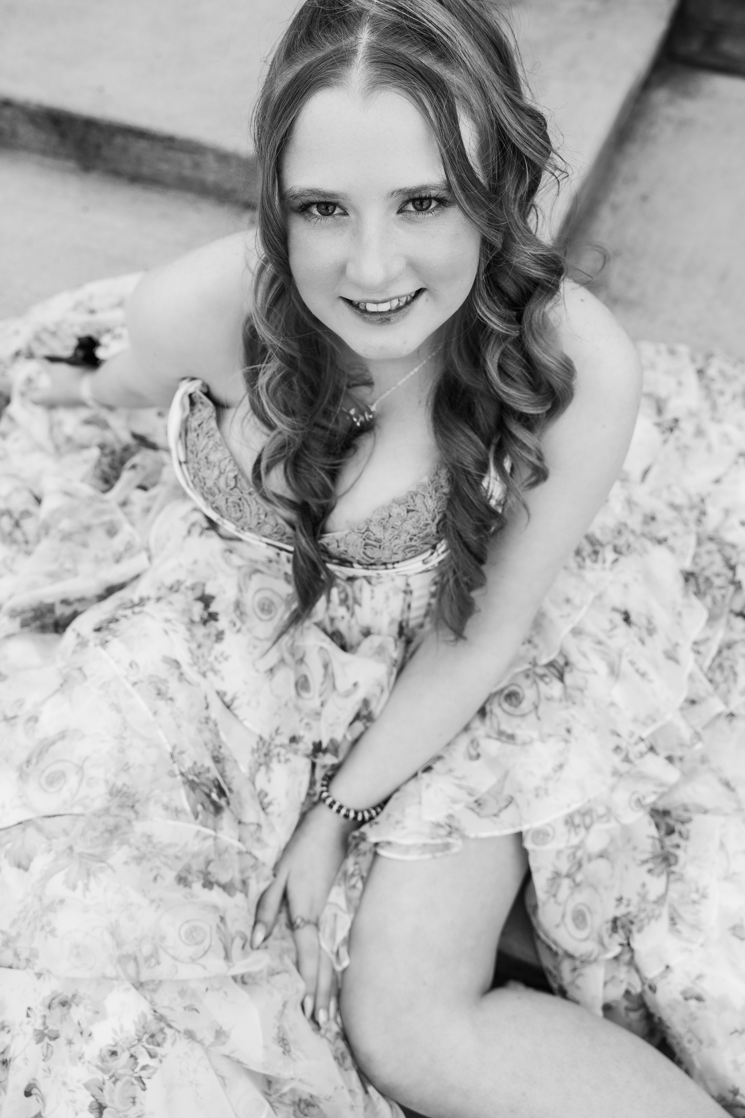 Black and white portrait of someone in a floral dress with long curled hair sitting on the floor.