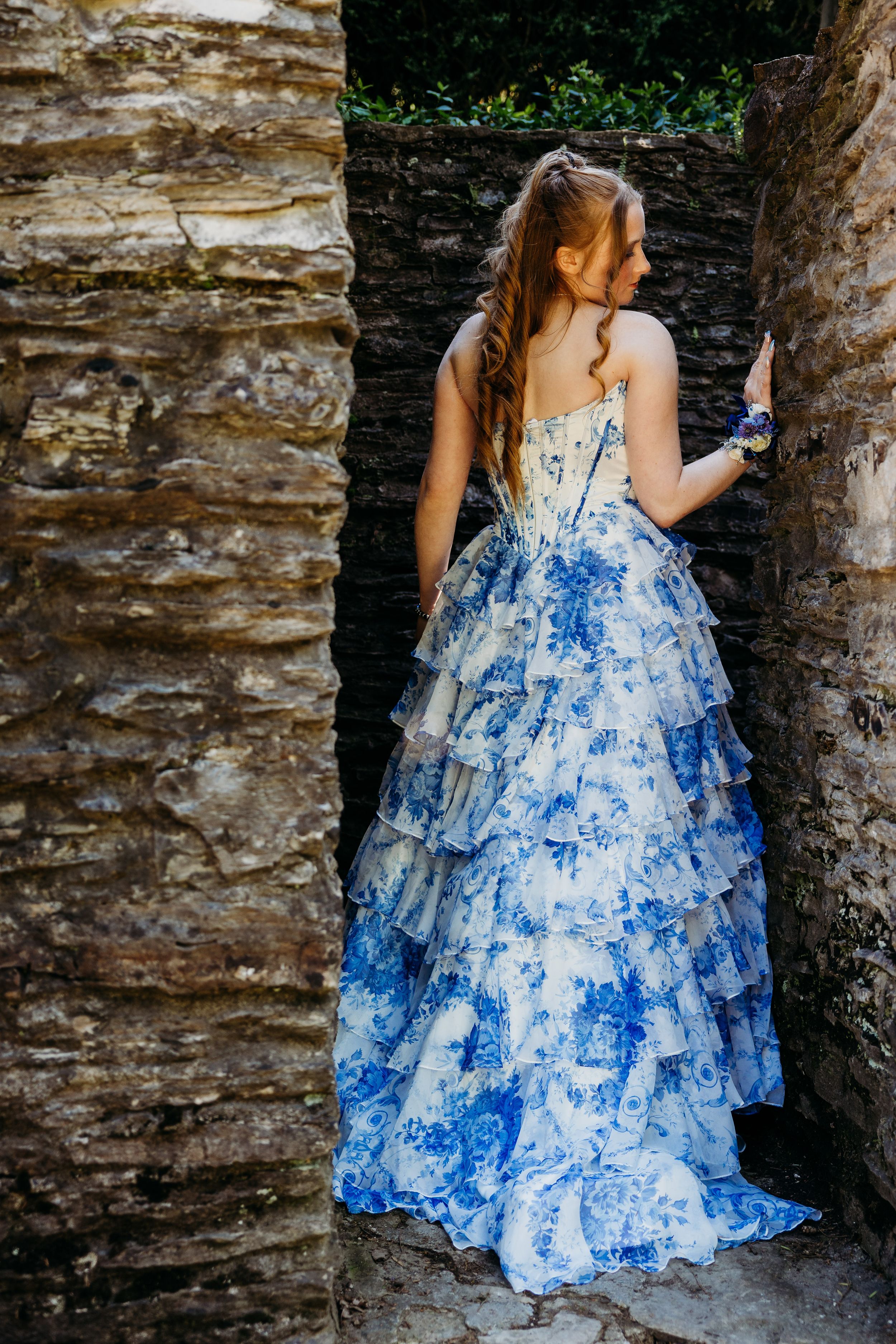 A woman in a flowing white and blue floral gown stands between stone walls in an elegant outdoor setting.