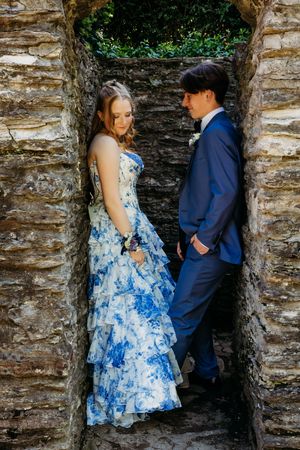 A couple in formal attire pose together in a stone alcove, with her wearing a blue floral gown and him in a navy suit.