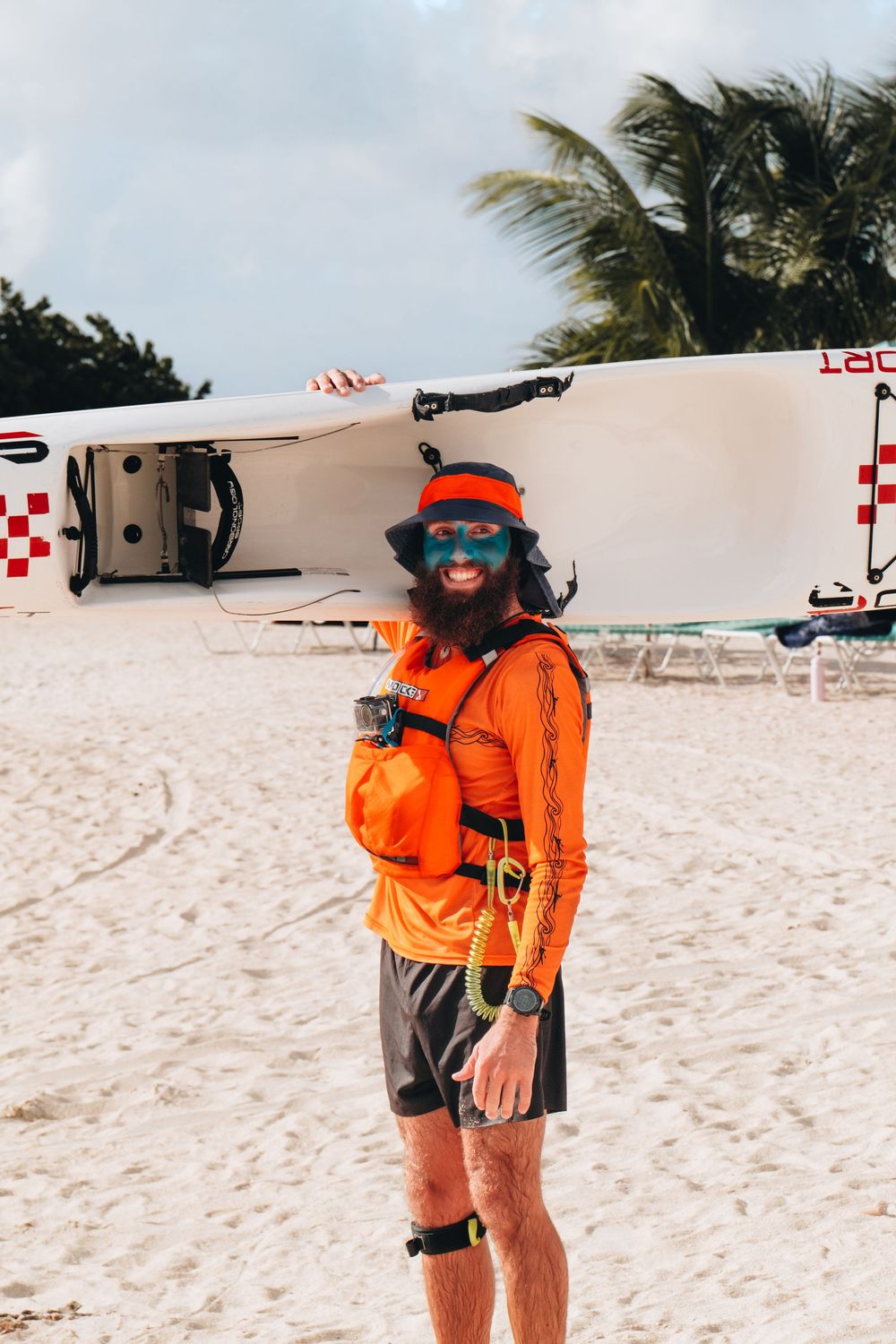 A beach lifeguard in bright orange safety gear stands on white sand with palm trees and water safety equipment.