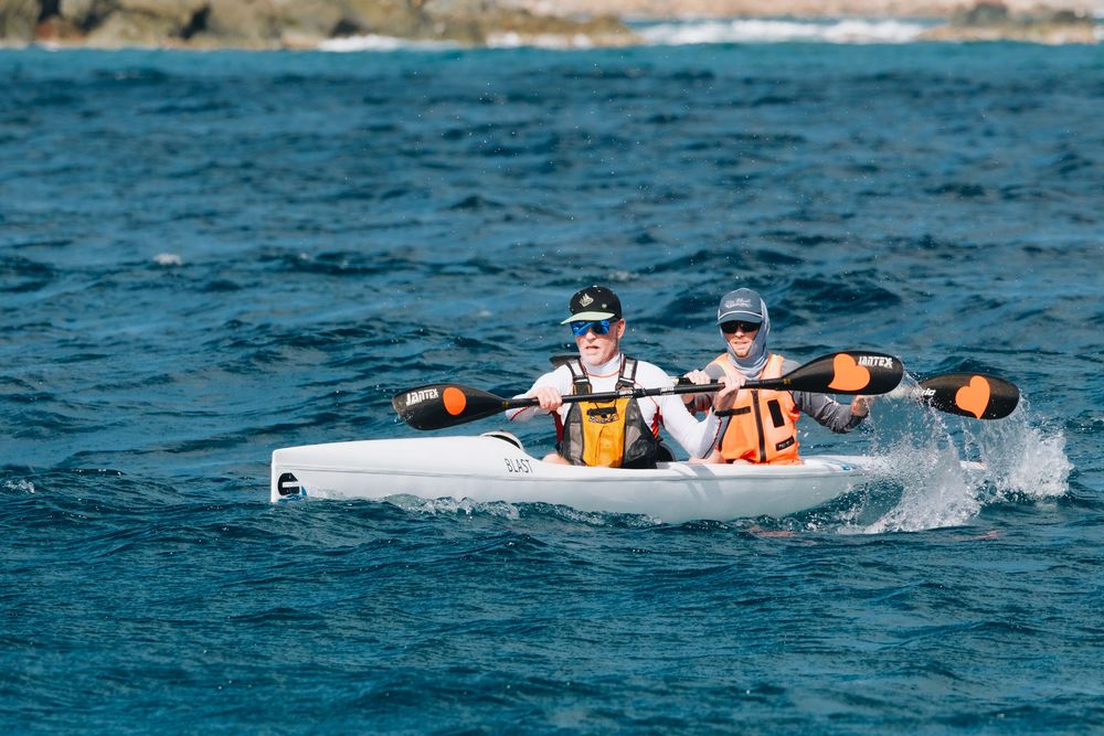 Two kayakers paddle through blue ocean waters in a white kayak near a rocky coastline on a sunny day.
