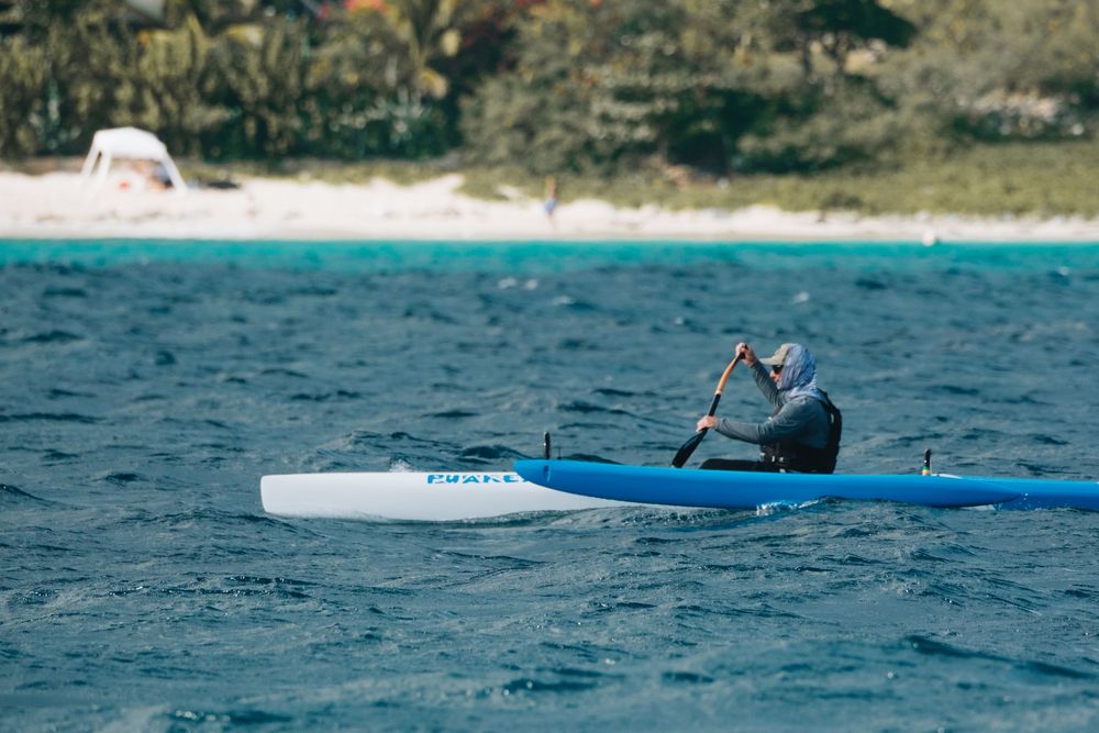 Kayaker paddles a blue and white kayak across turquoise ocean waters near a scenic beach.