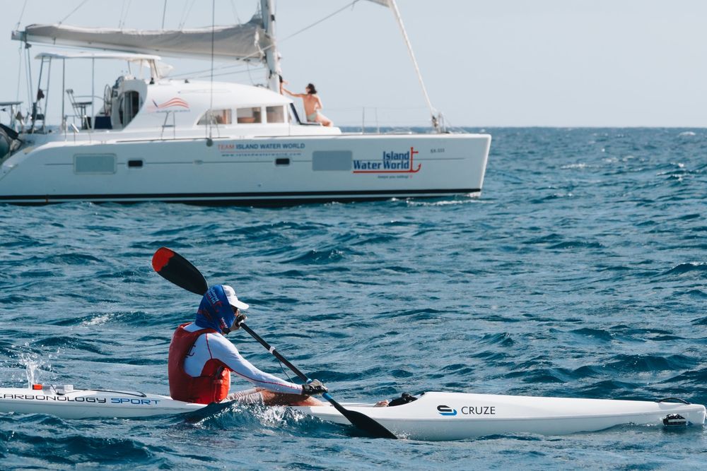 Sea kayaker paddles past a white catamaran yacht anchored in beautiful blue ocean waters.
