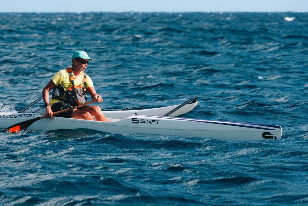 A paddler navigates choppy blue ocean waters on a sleek white surf ski in warm weather conditions.
