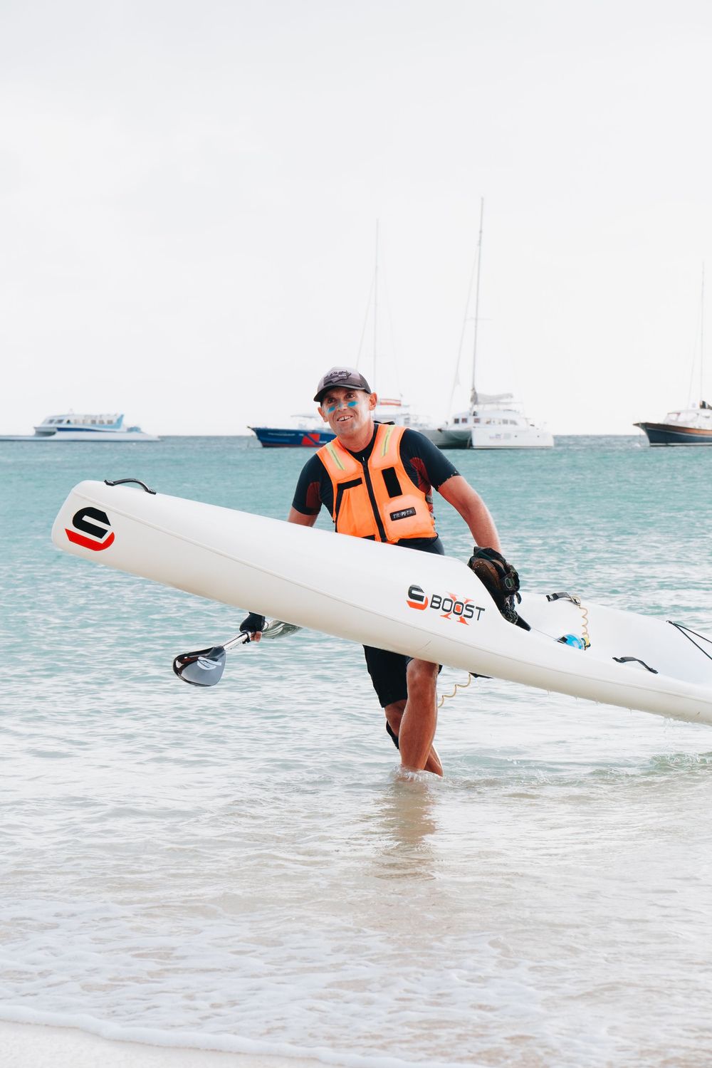 Person carrying a white surfboard wearing an orange life vest walks along a tropical beach with boats in background.