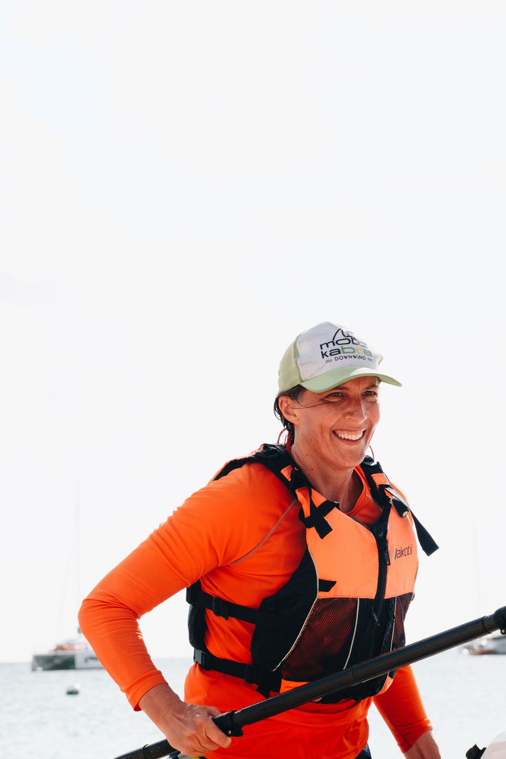 An adventurer in orange athletic gear and a baseball cap smiles while carrying winter sports equipment in a snowy setting.