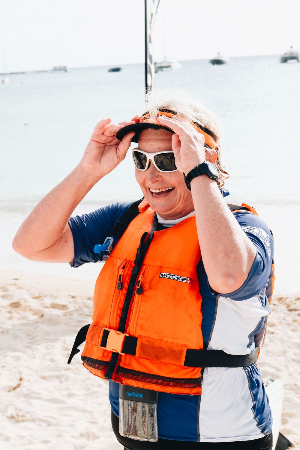 Person wearing orange life vest and sunglasses smiles while adjusting shades on sunny beach.