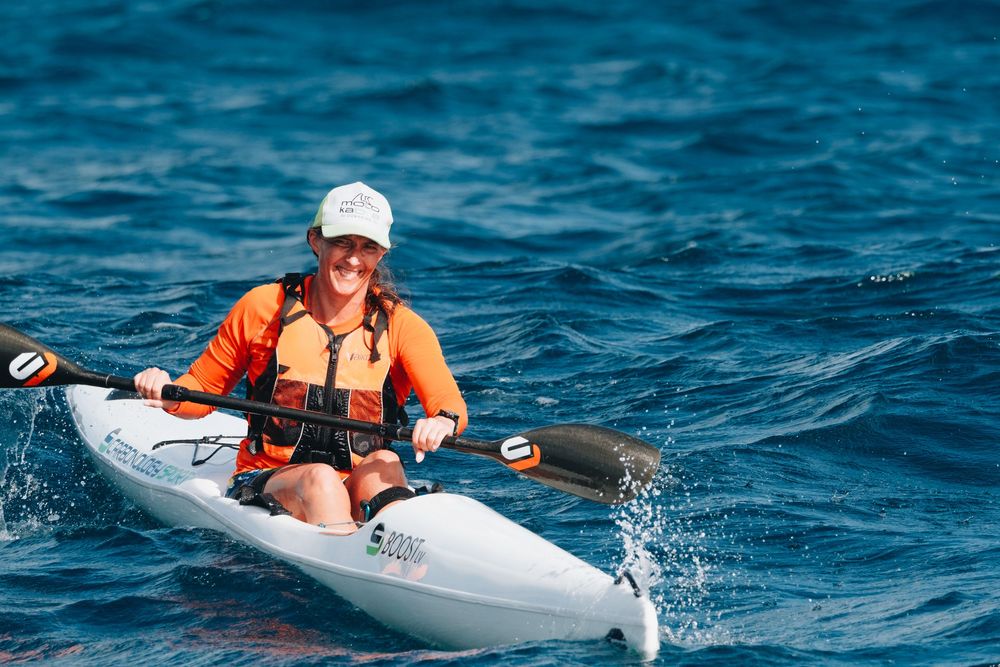 A kayaker in an orange life vest paddles a white kayak across bright blue waters on a sunny day.